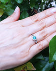 Hand wearing a ring with a round cut emerald against a leafy background.
