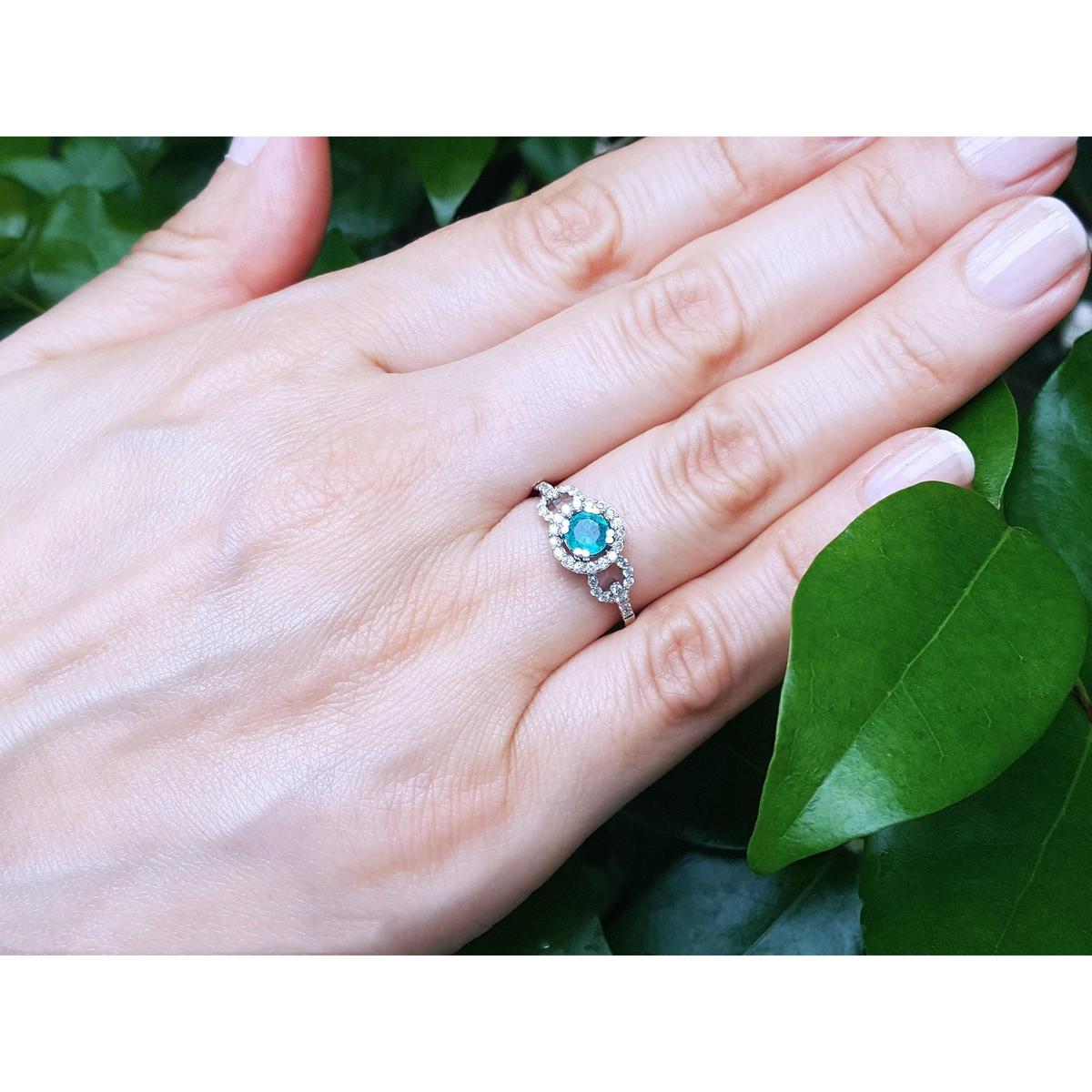 Hand wearing a white gold ring with a round cut emerald against a green leafy background.