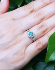 Hand wearing a white gold ring with a round cut emerald against a green leafy background.