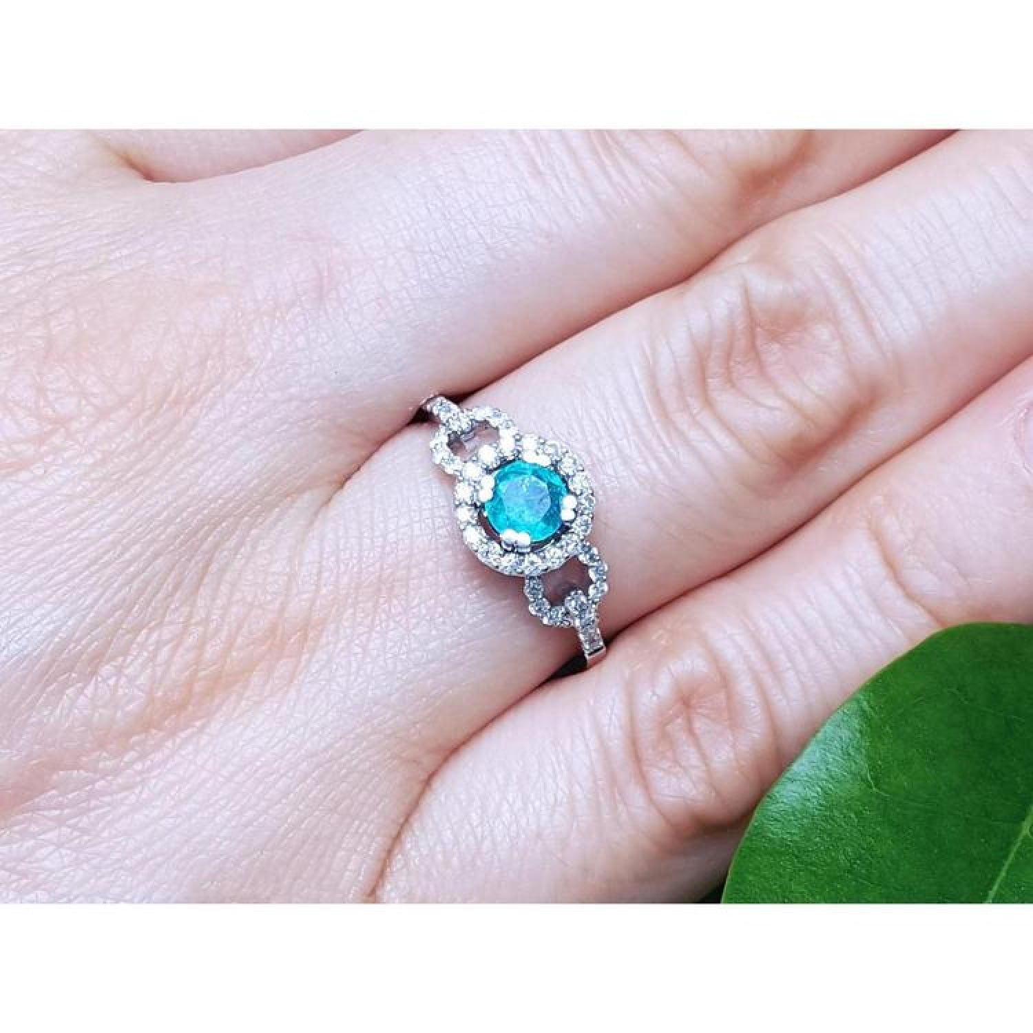 14K white gold ring with a round bluish-green emerald haloed by diamonds on a finger.