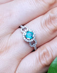 A white gold with a round emerald ring on a hand, with a green leaf in the background.