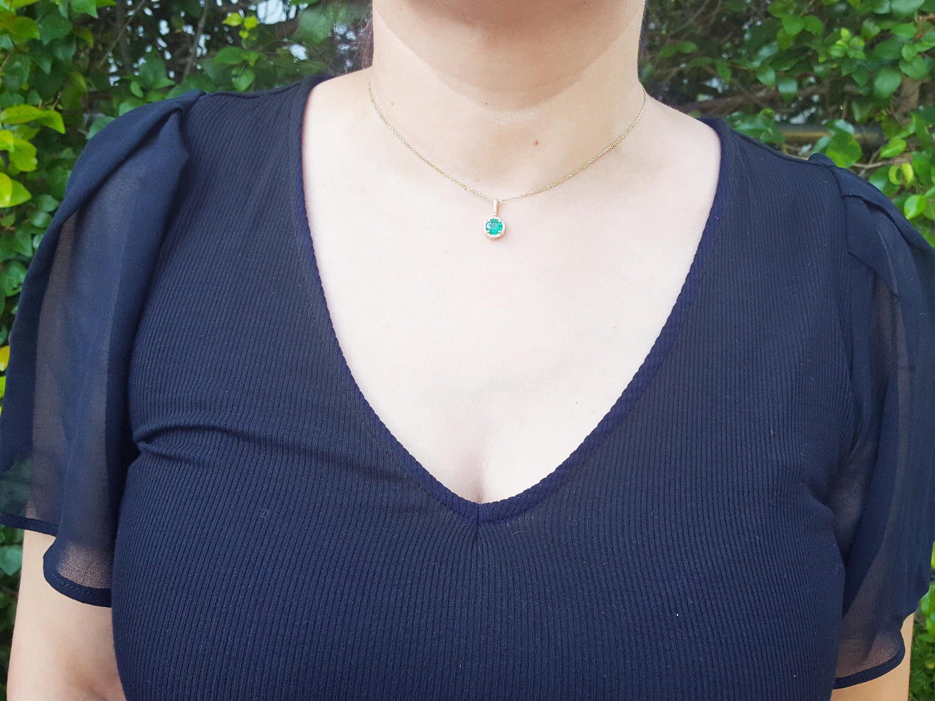 A woman wearing an emerald pendant necklace in a navy blue dress against a green leafy background.