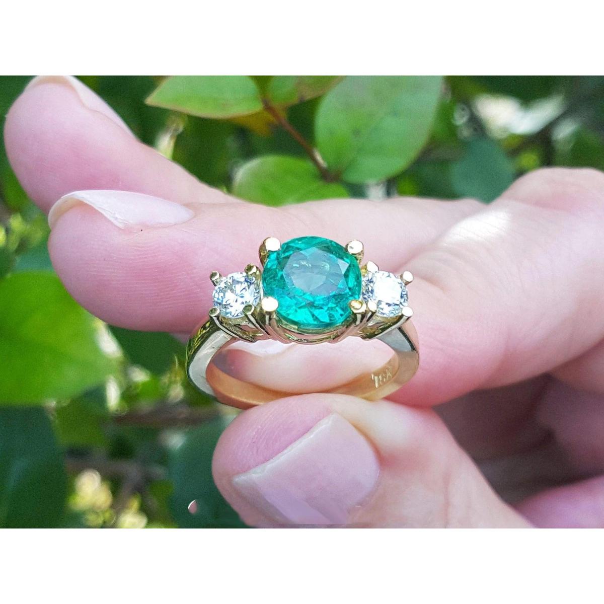 A 18k yellow gold ring with a round cut emerald held between fingers against a blurred green leafy background.