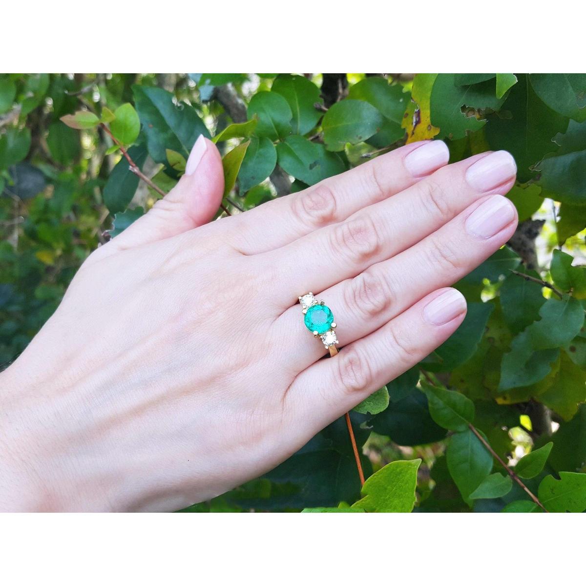 Hand wearing a round cut emerald and side diamonds ring against a green leafy background.