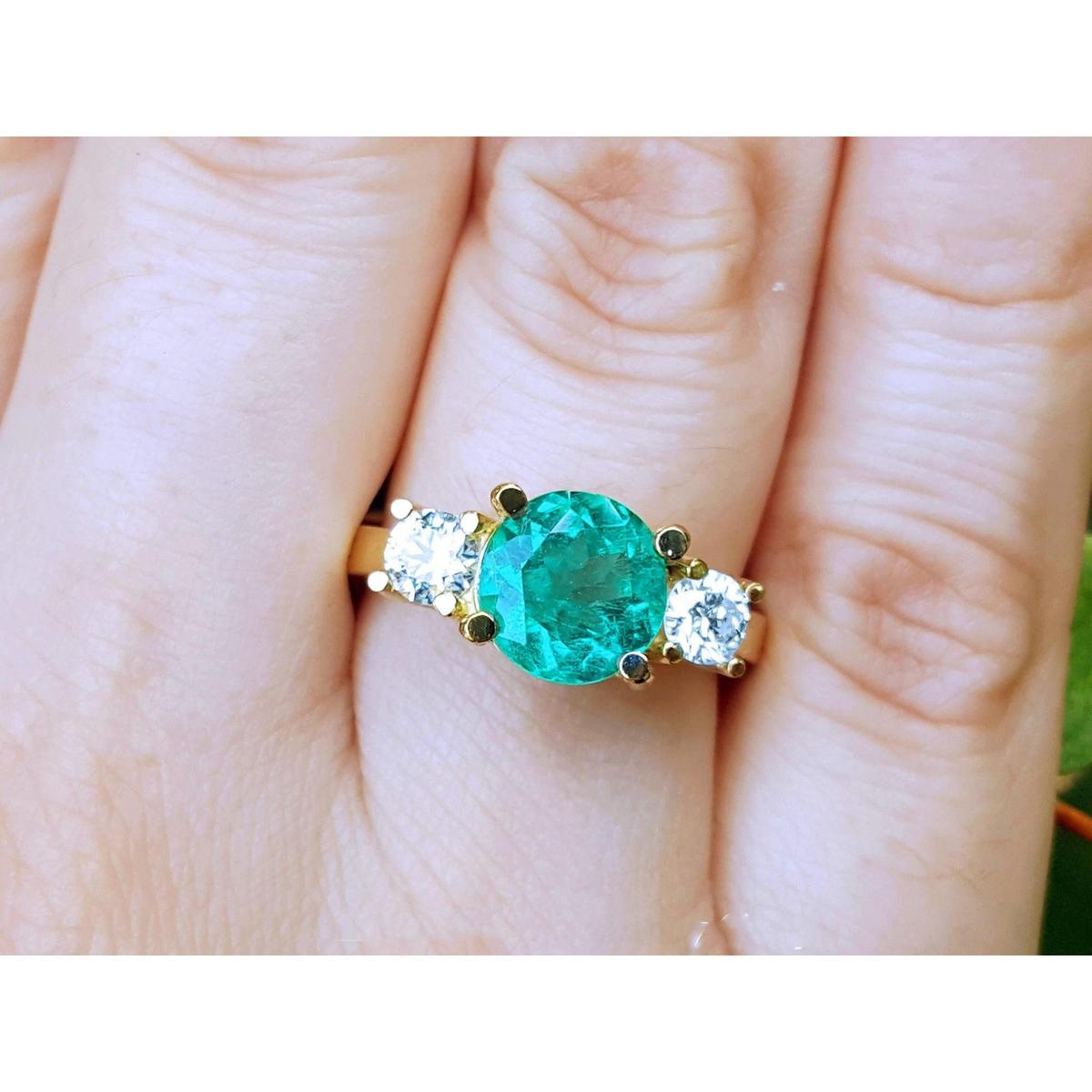 18k yellow gold round cut emerald ring with two side diamonds on a finger.