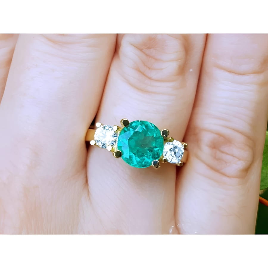 18k yellow gold GIA certified round cut colombian emerald ring with two side diamonds on a finger.