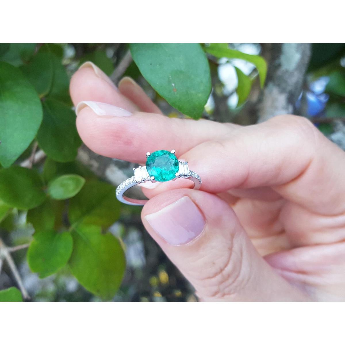 Hand holding a ring with a round cut emerald against a blurred natural background.