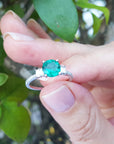 Hand holding a ring with a round cut emerald against a blurred natural background.