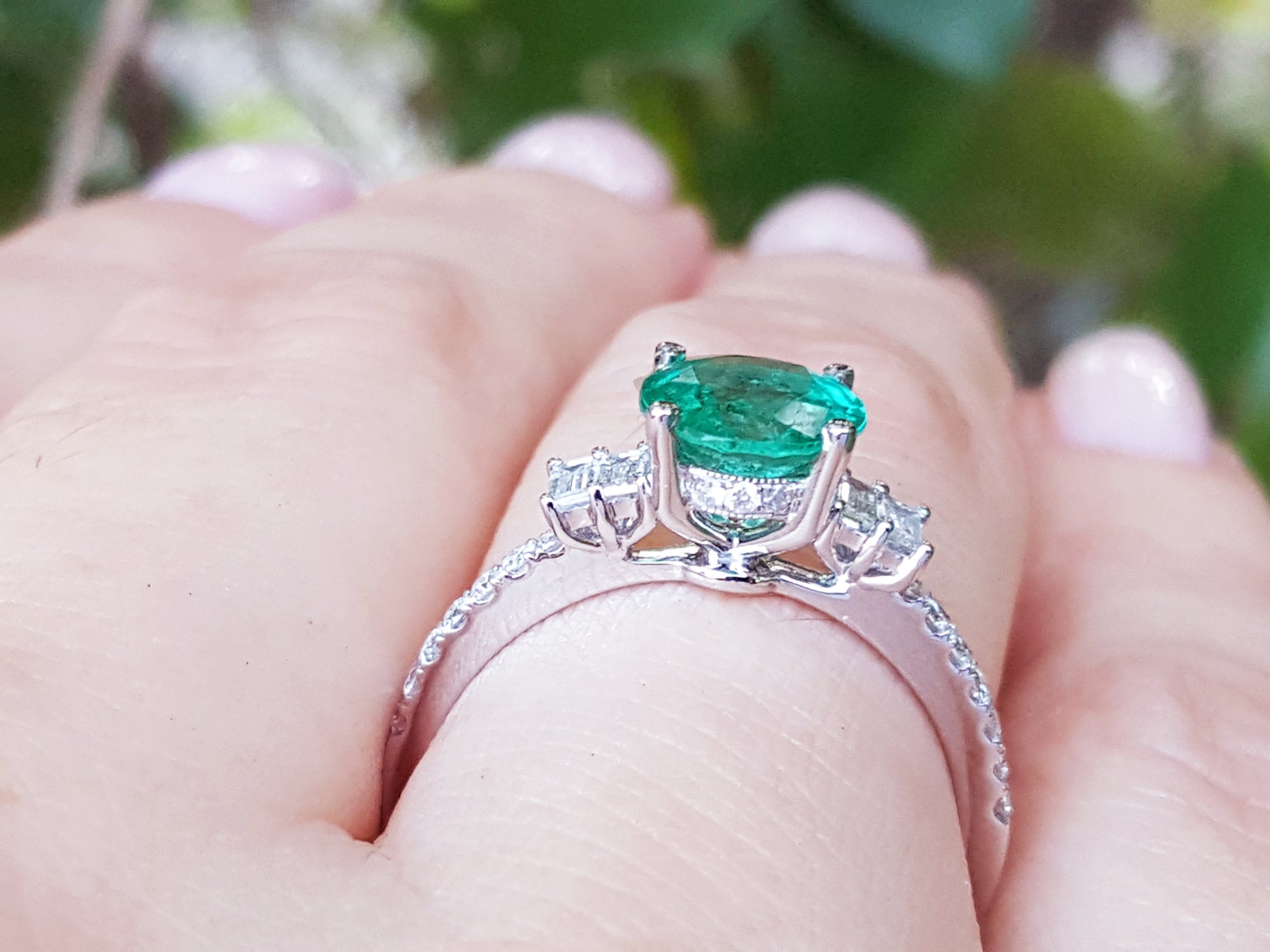 Emerald ring with diamond accents on a hand against a blurred green background.