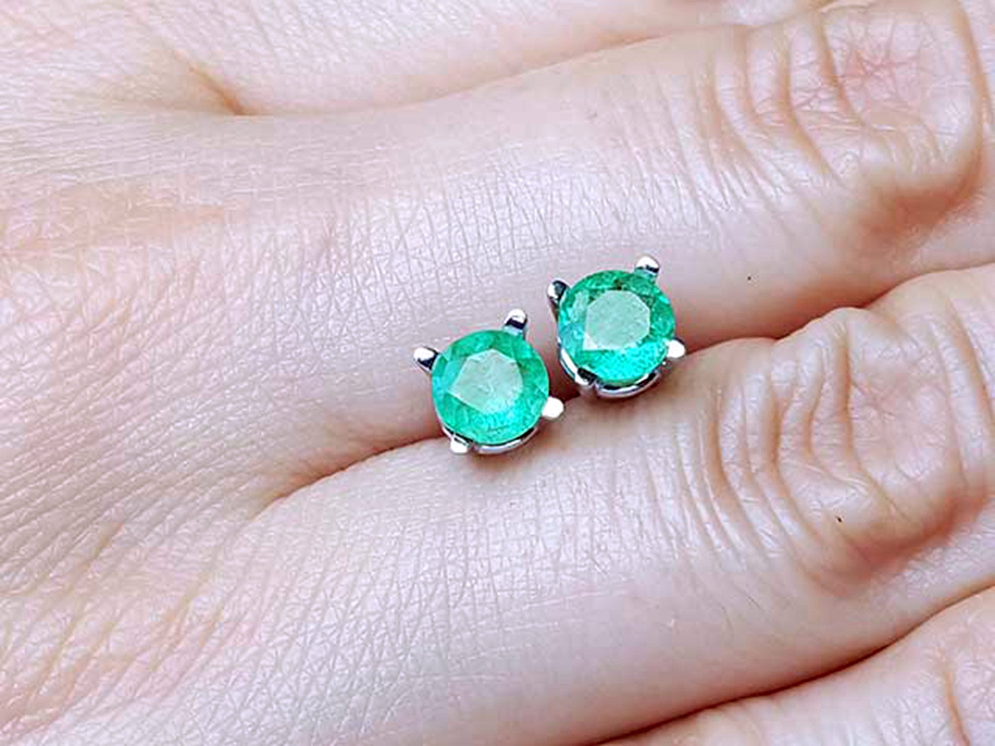 Pair of round cut emerald stud earrings between fingers.
