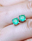 Pair of round cut emerald stud earrings between fingers.