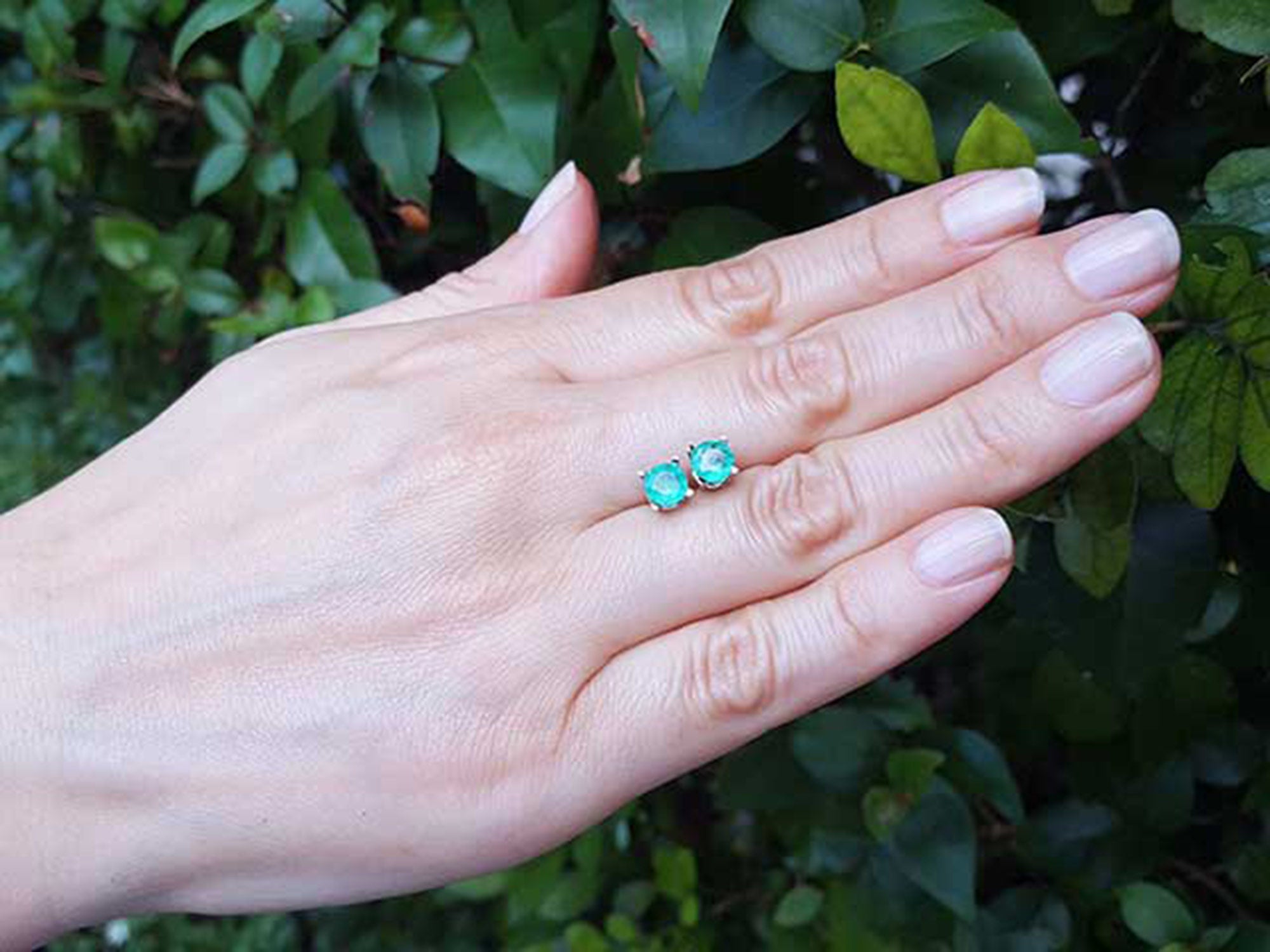 Hand displaying pair of emerald stud earrings round cut against a green leafy background.