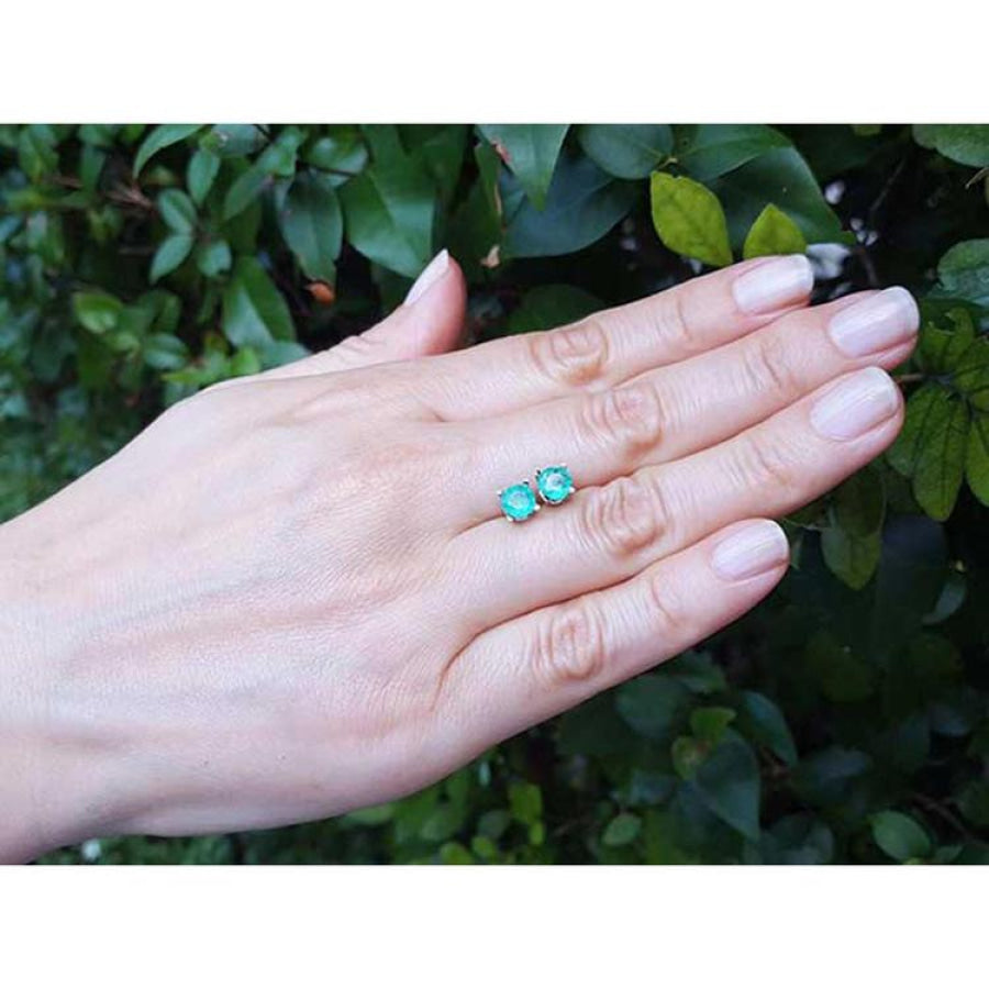 Round Colombian emerald stud earrings in 14k white gold, displayed on a hand against green foliage.