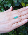 Hand displaying pair of emerald stud earrings round cut against a green leafy background.