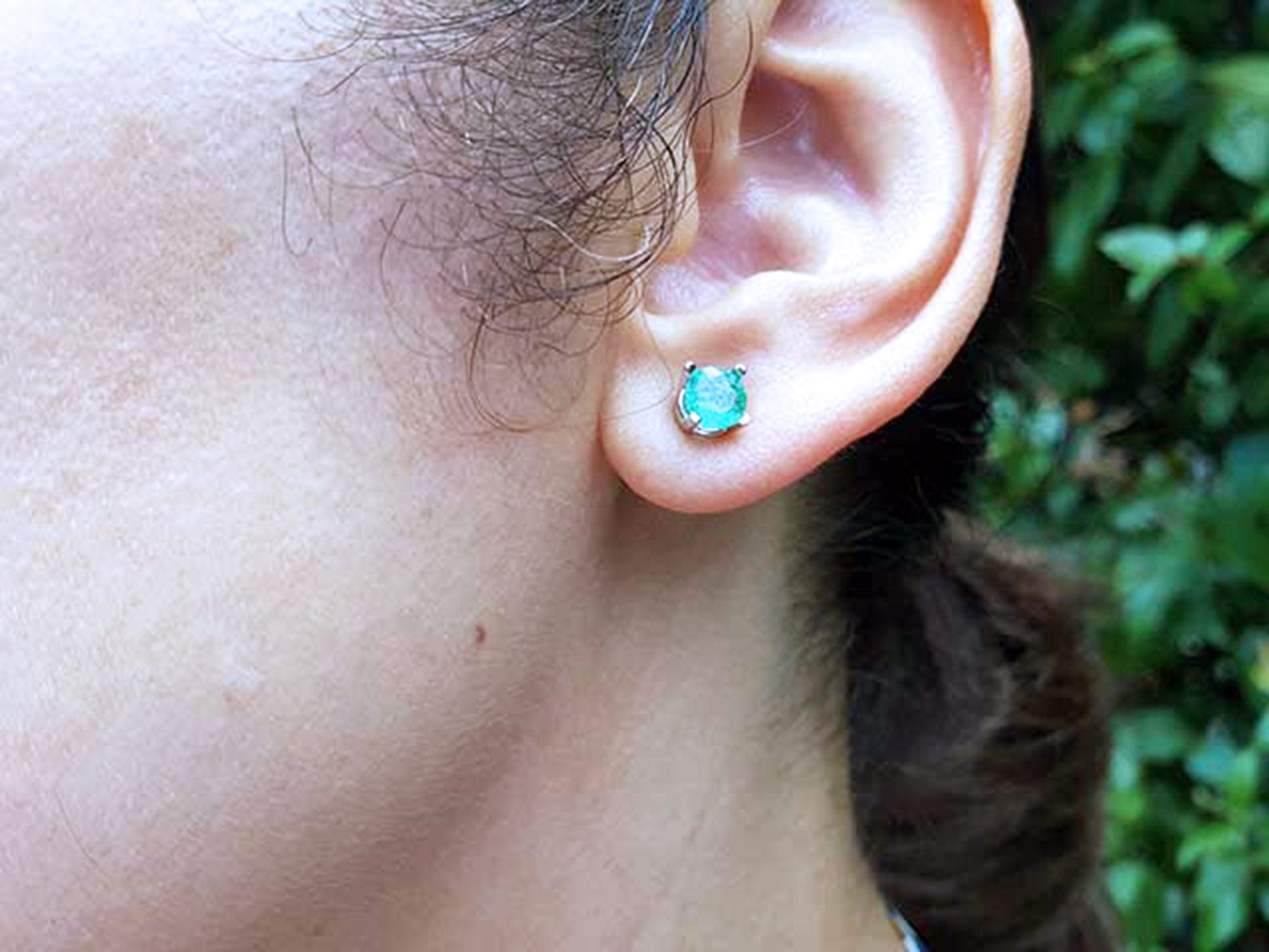 Close-up of a woman&#39;s ear with an emerald stud earring against a blurred natural background.
