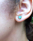 Close-up of a woman's ear with an emerald stud earring against a blurred natural background.