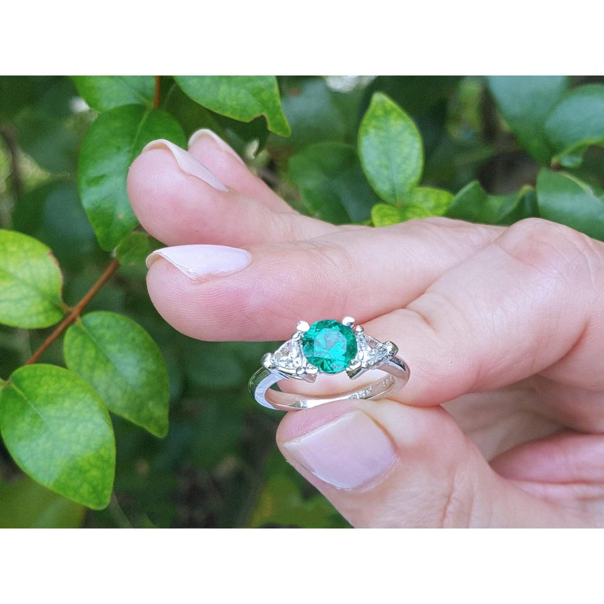Hand holding a brides' emerald engagement rings against a green leafy background.