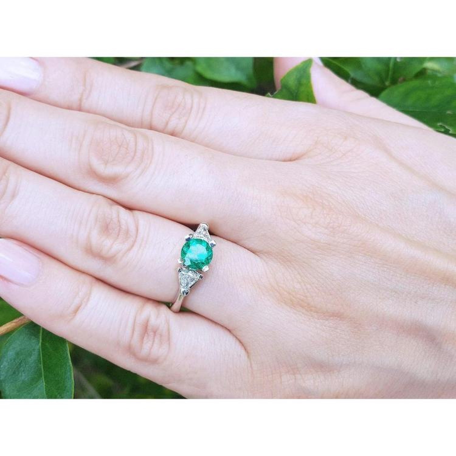 1.00 ct. round emerald ring with trillion side diamonds on a silver band, worn on a hand.