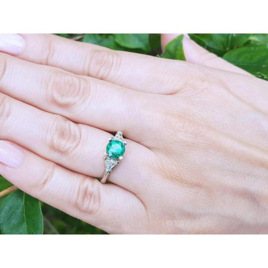 1.00 ct. round emerald ring with trillion side diamonds on a silver band, worn on a hand.