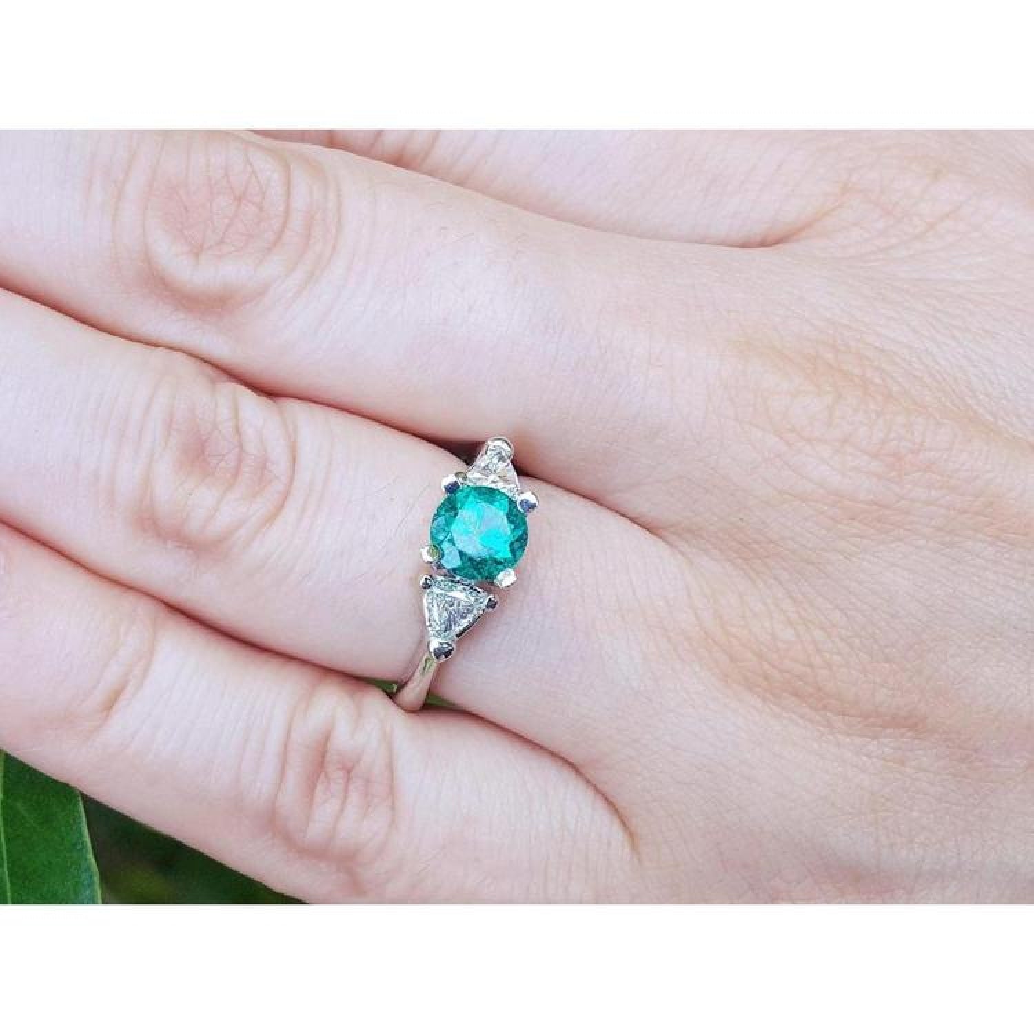 1.00 ct. round emerald ring with trillion side diamonds on a 14K white gold band, worn on a finger.