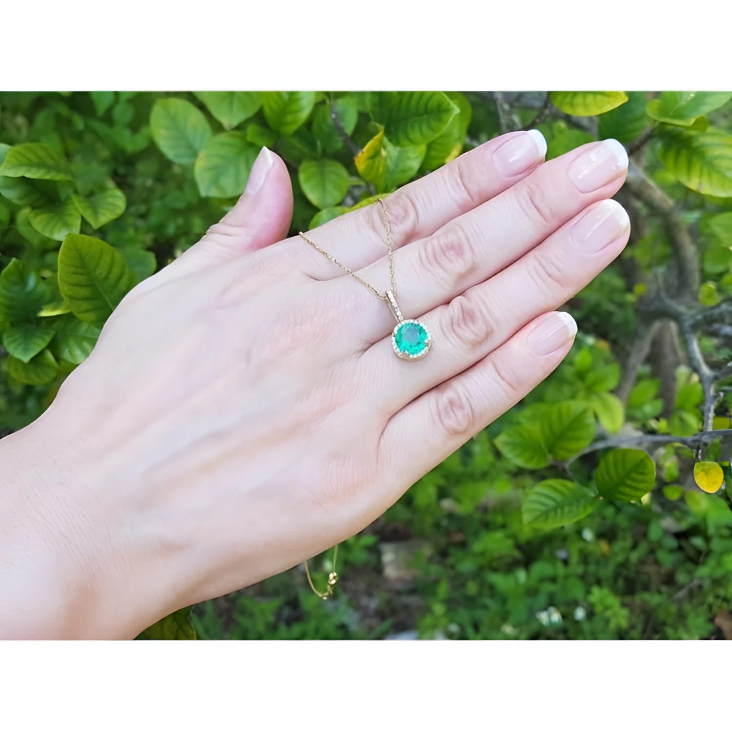 Round-cut natural emerald pendant necklace with halo diamonds in 14K yellow gold, worn on a hand.