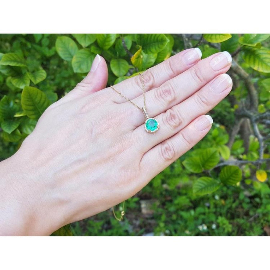 Round-cut natural emerald pendant necklace with halo diamonds in 14K yellow gold, worn on a hand.