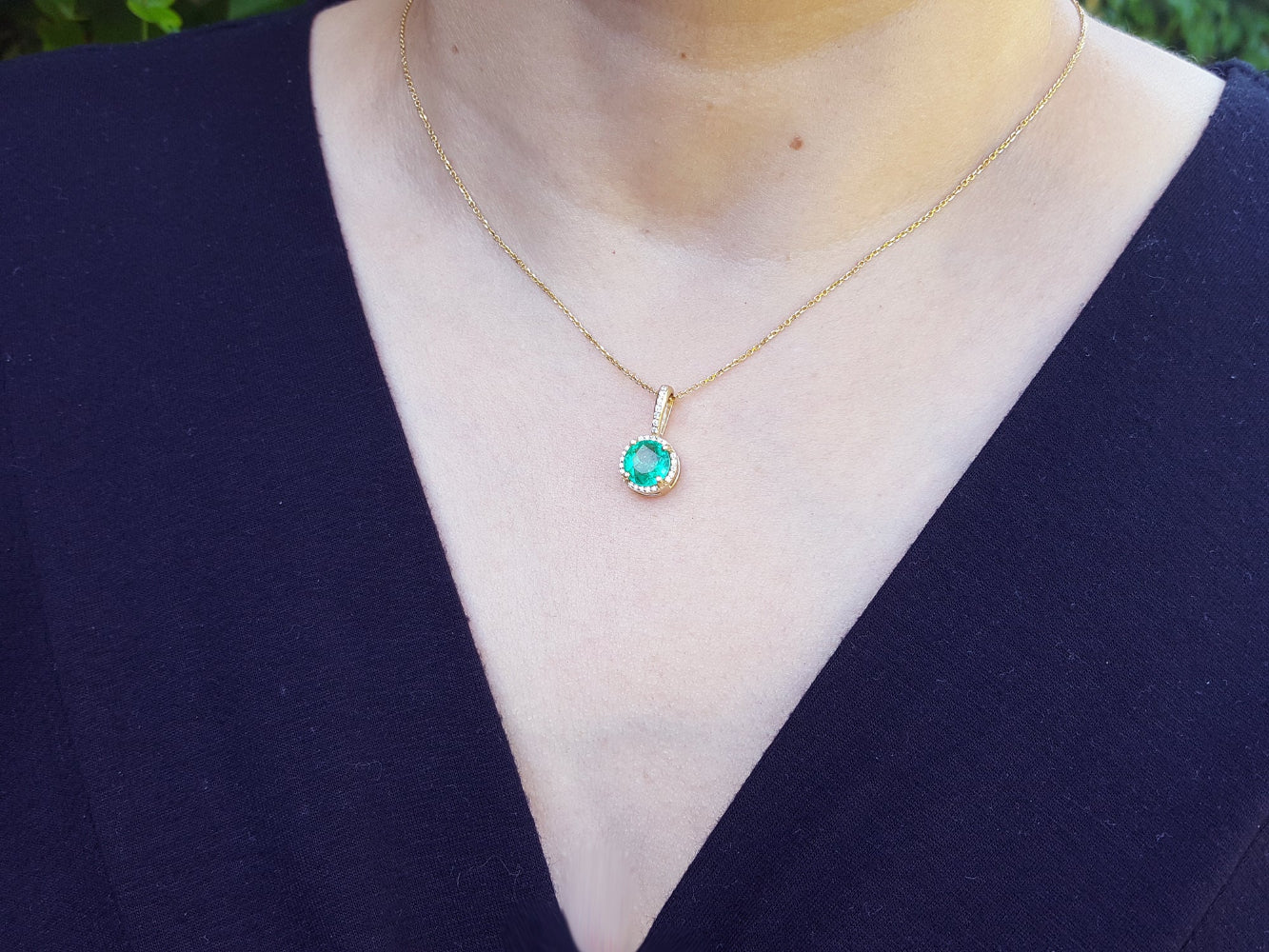 Gold necklace with a round cut emerald pendant on a woman&#39;s neck wearing a dark blue top.