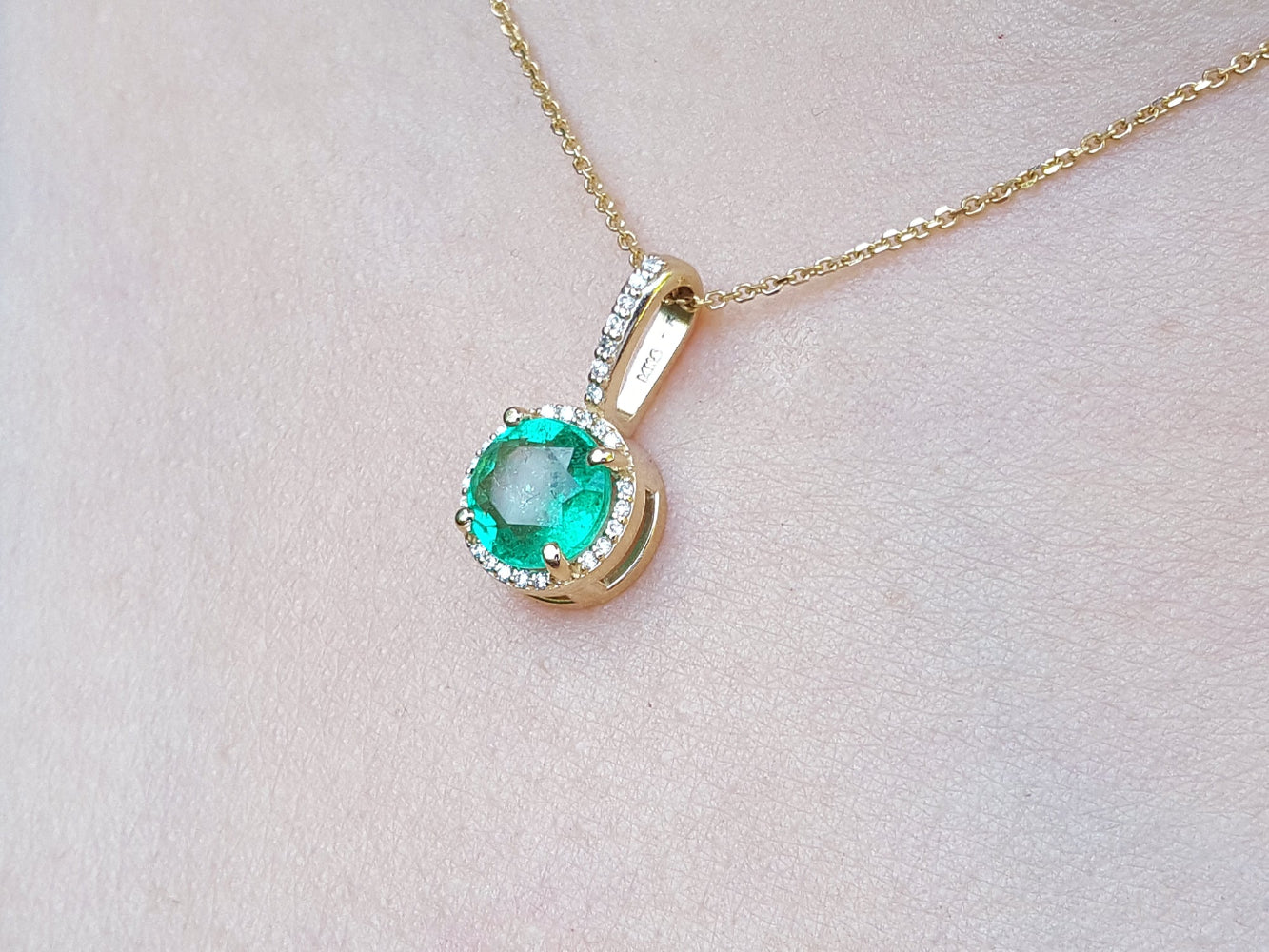 Gold necklace with a round cut natural colombian emerald pendant on a light pink skin background,