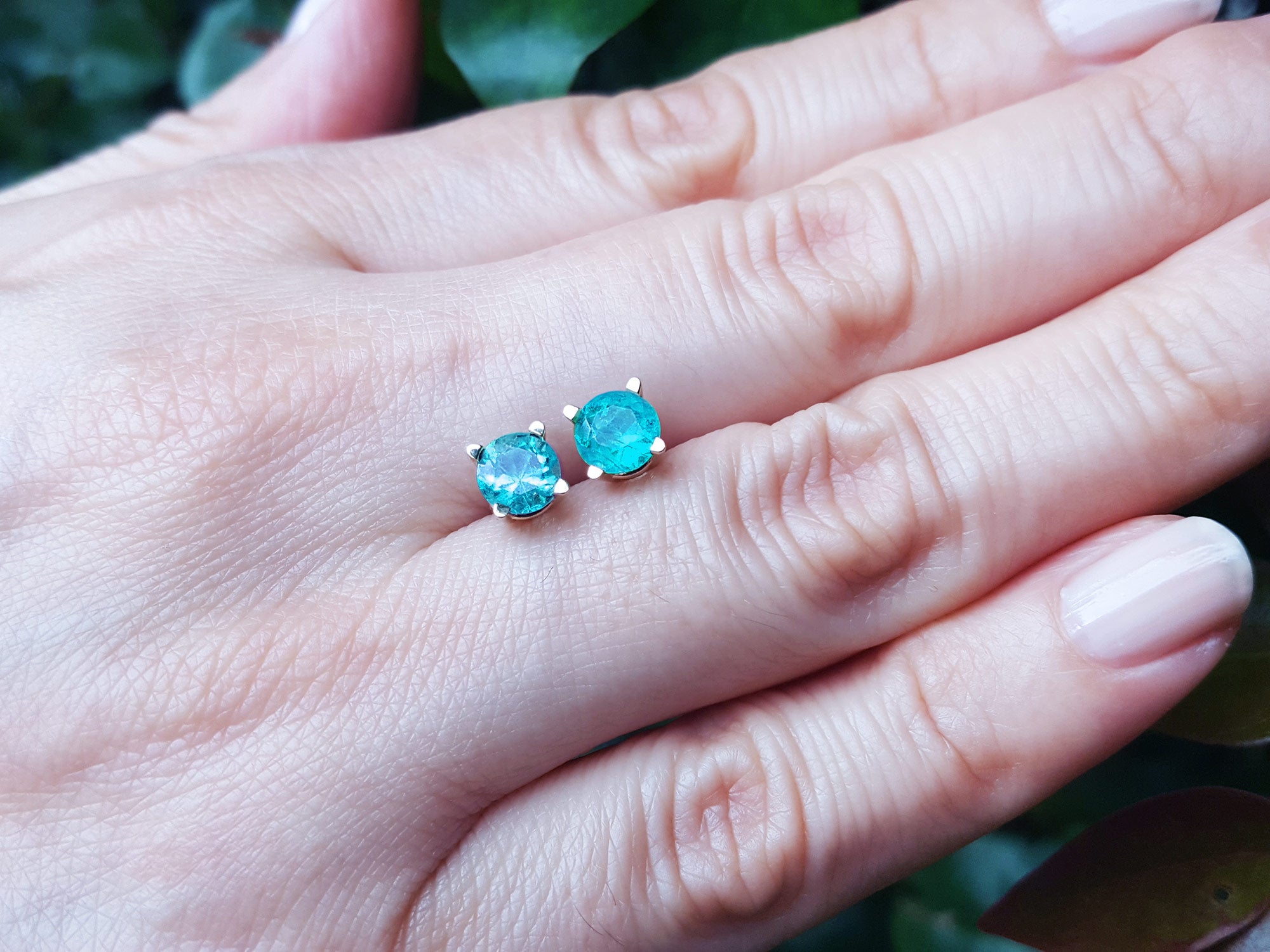 Hand between fingers holding a 14k yellow gold emerald stud earrings round cut against a natural background.