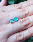Hand between fingers holding a 14k yellow gold emerald stud earrings round cut against a natural background.
