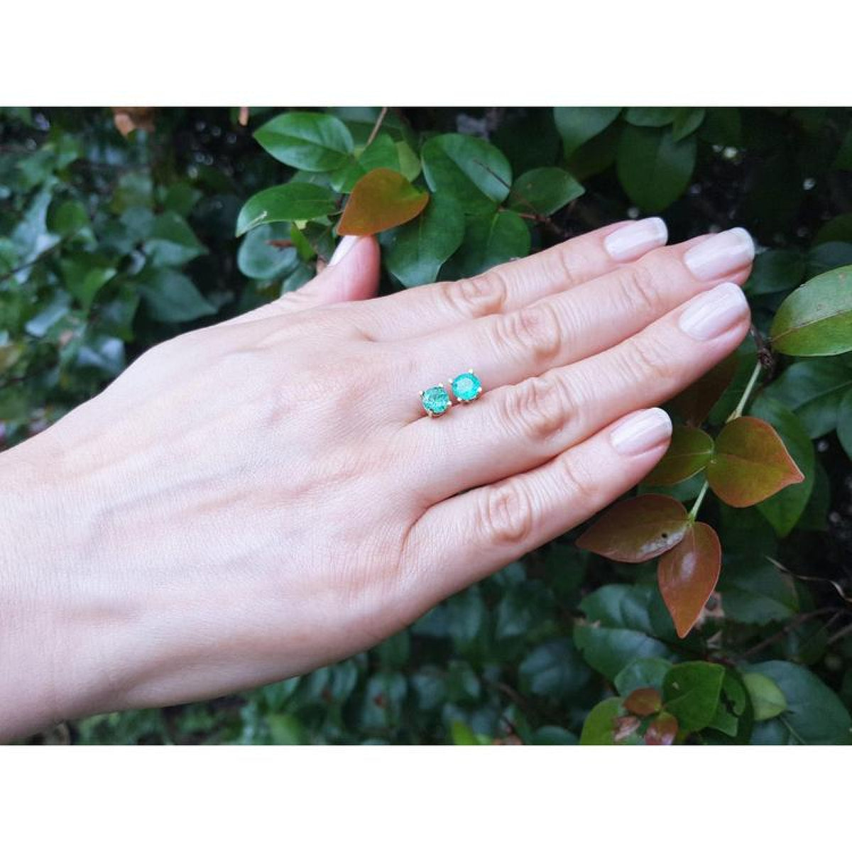 Round natural Colombian emerald stud earrings in 14K gold, displayed on a hand against green foliage.