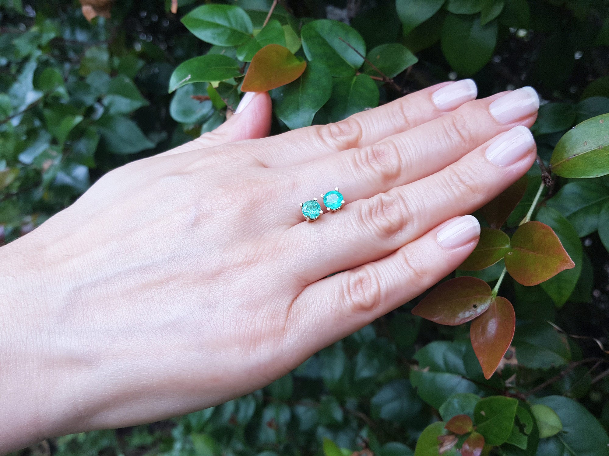 Hand showing a pair of round cut emerald stud earrings against a green leafy background.