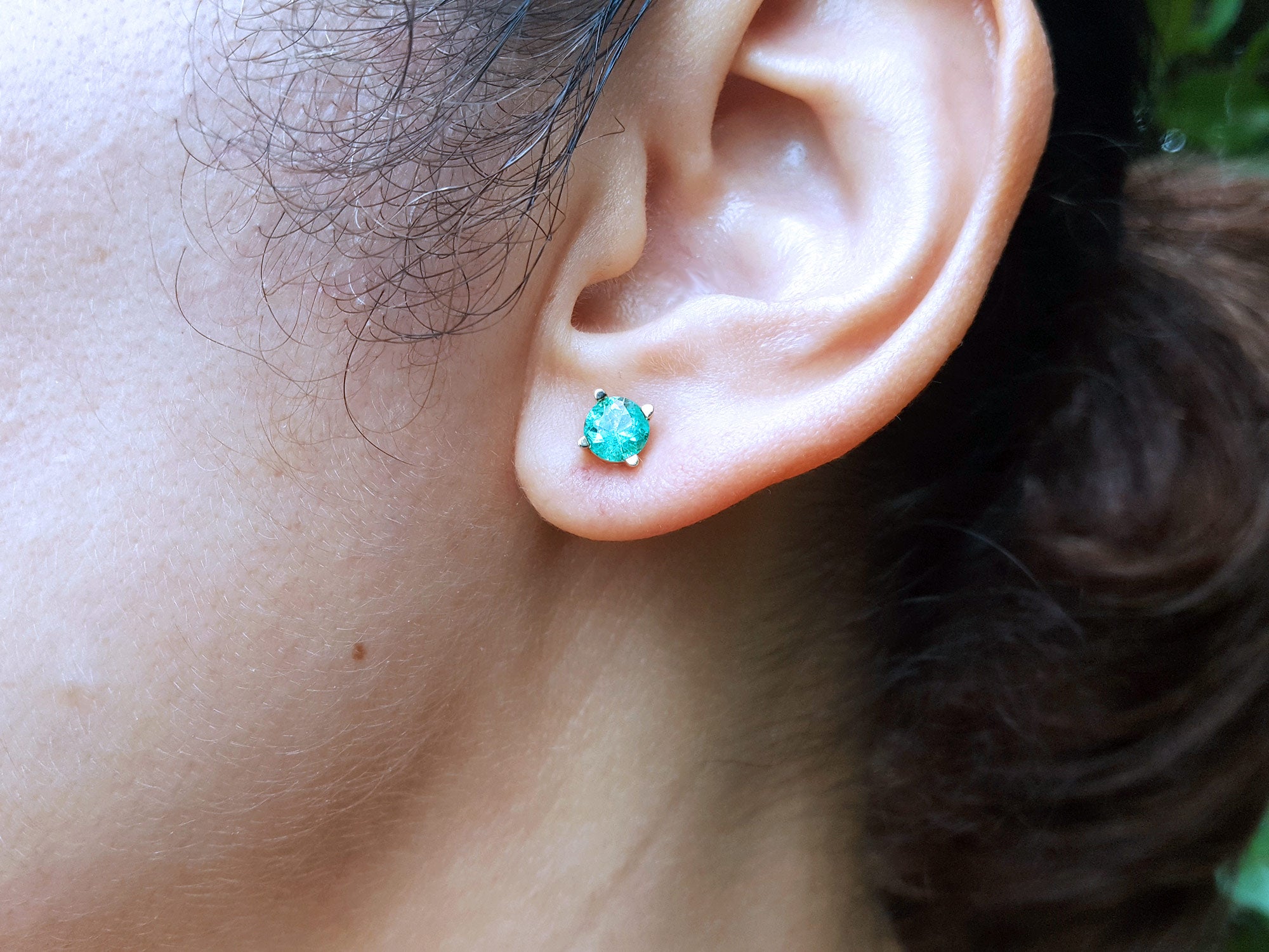 Close-up of a woman&#39;s ear wearing a 14k yellow gold emerald round cut stud earring.