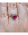 A gold ring with a large heart-shaped pink gemstone and a smaller clear heart-shaped gemstone, shown worn on a person's finger.