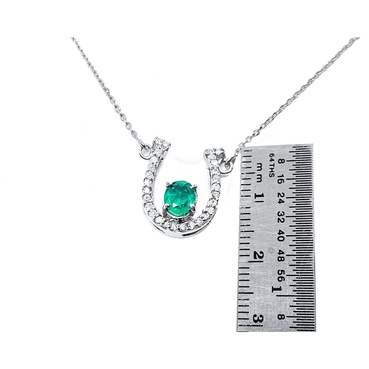Horseshoe-shaped pendant with natural emerald gemstone and diamonds next to a ruler for scale on a white background