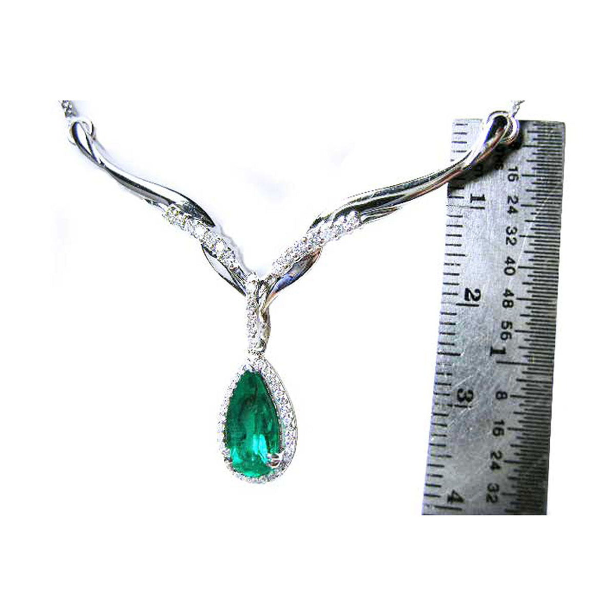 White gold necklace with a pear cut emerald gemstone in a halo diamond next to a ruler on a white background