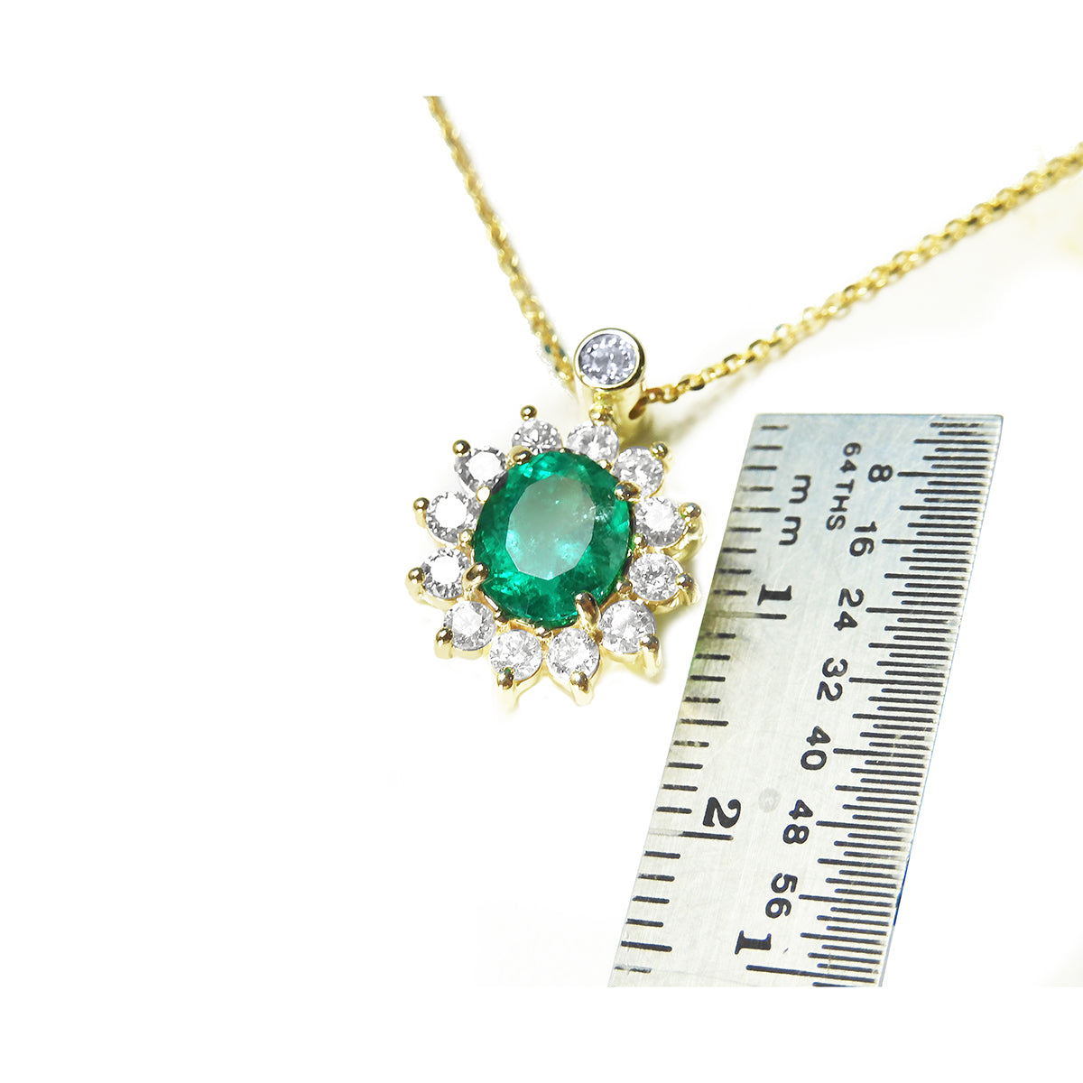 A yellow gold necklace with an oval emerald pendant next to a ruler for scale on a white background
