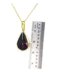 Teardrop-shaped pendant with gold chain and embedded red and green black opal, next to a ruler for scale.
