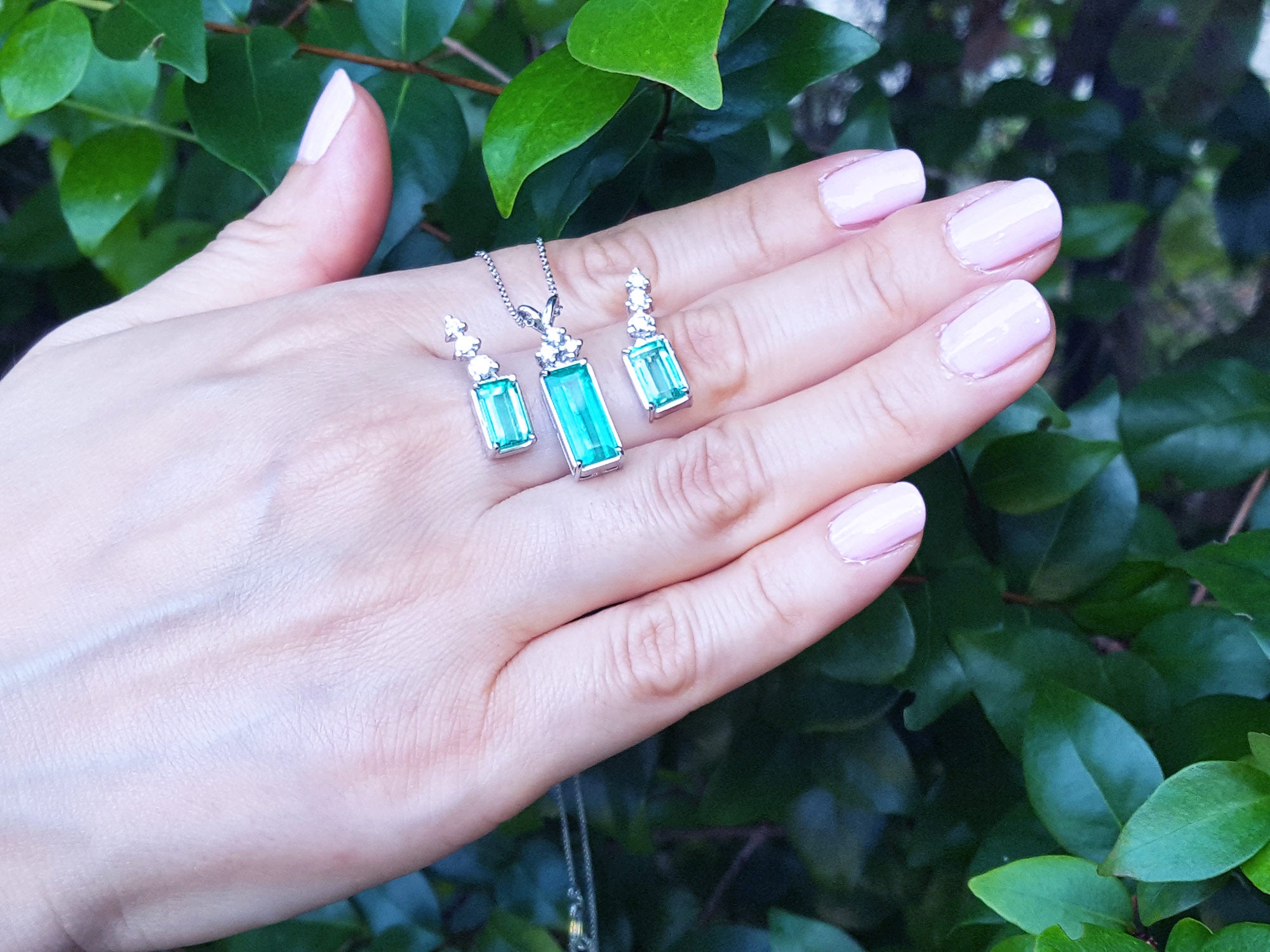 A hand displaying a trio set of emerald earrings and pendant against a green leafy background