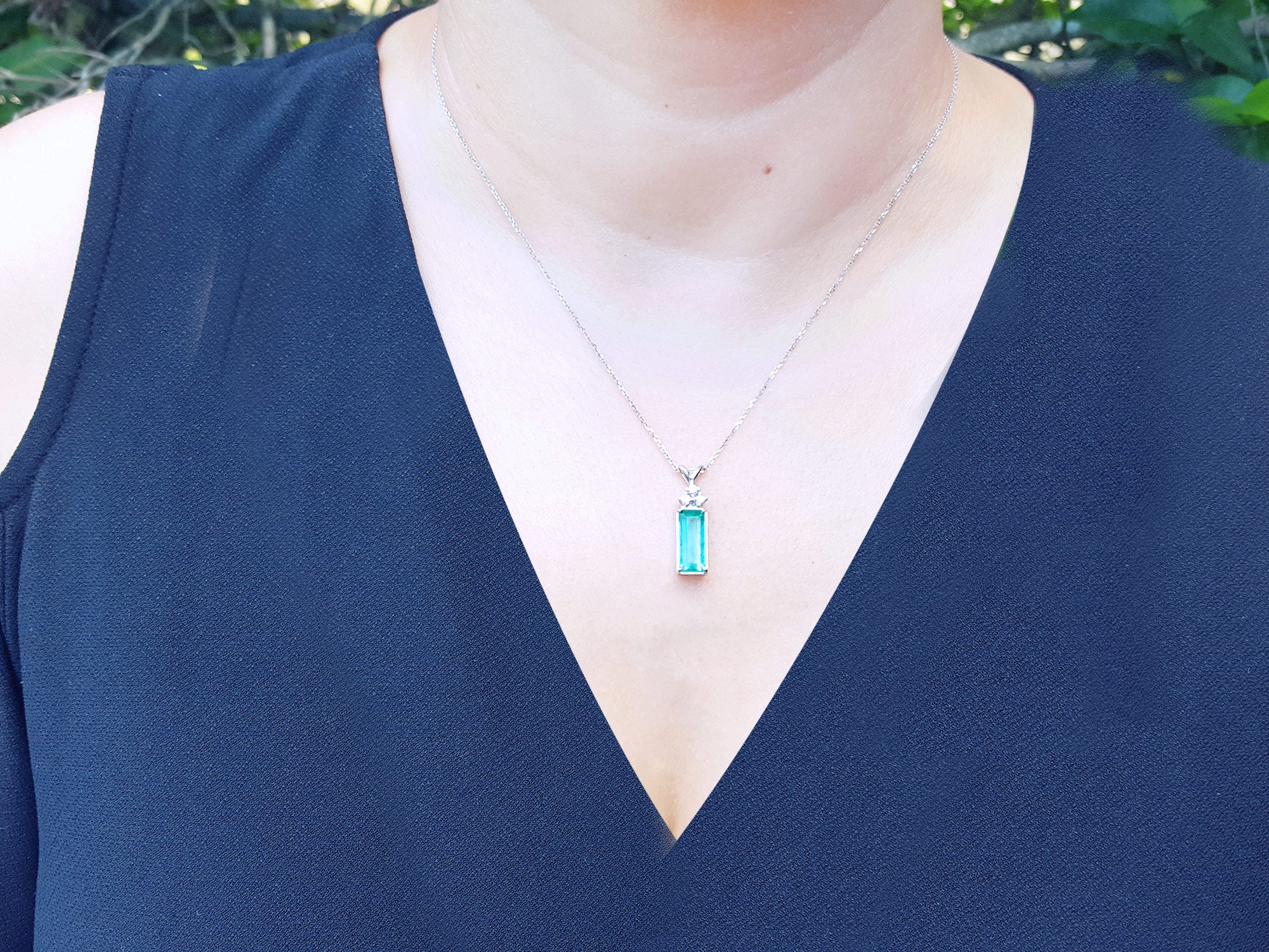 A woman wearing a blue top with a necklace featuring a rectangular emerald pendant.
