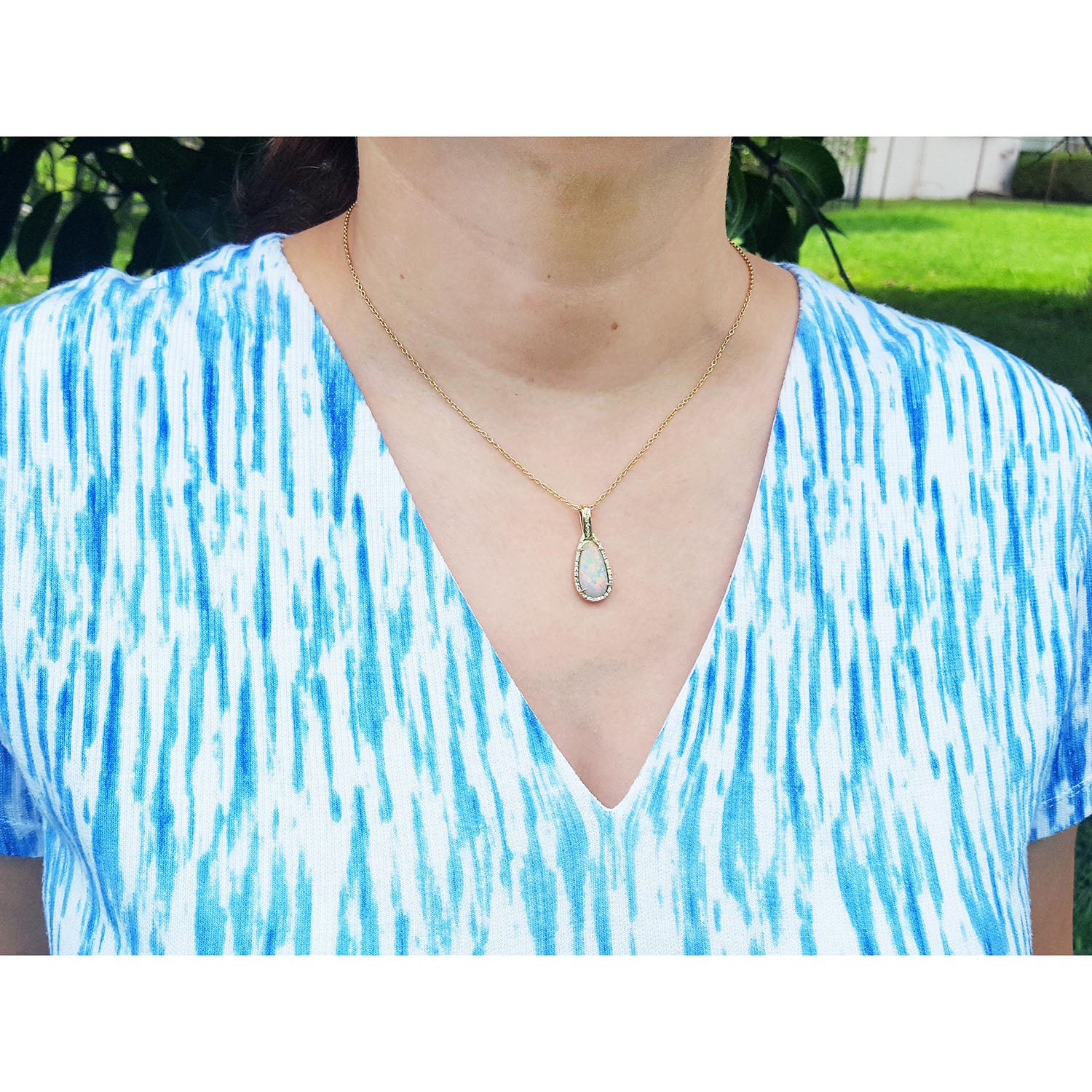 Person wearing a blue and white patterned top with a gold necklace featuring an opal pendant.