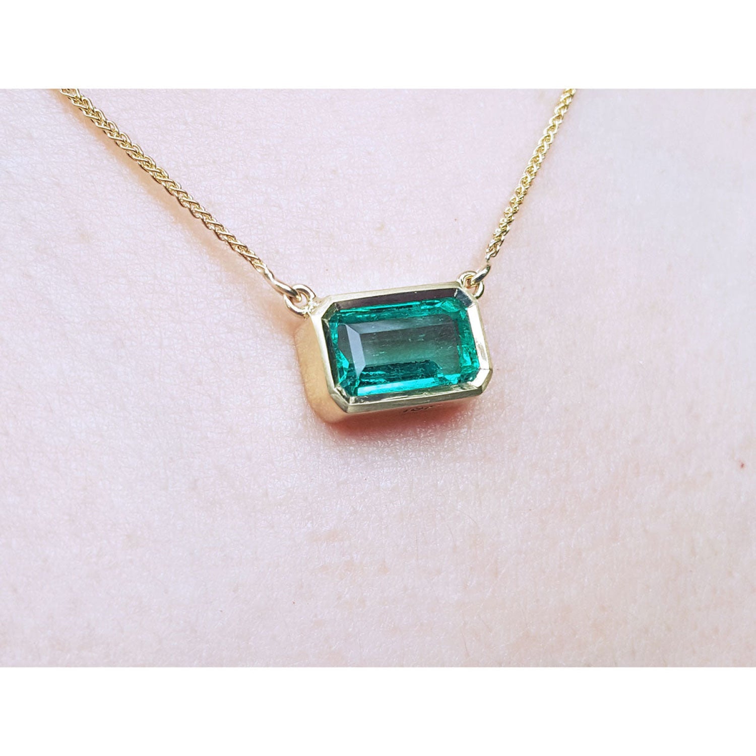 East-west emerald-cut solitaire emerald necklace in 18K yellow gold, bezel set.