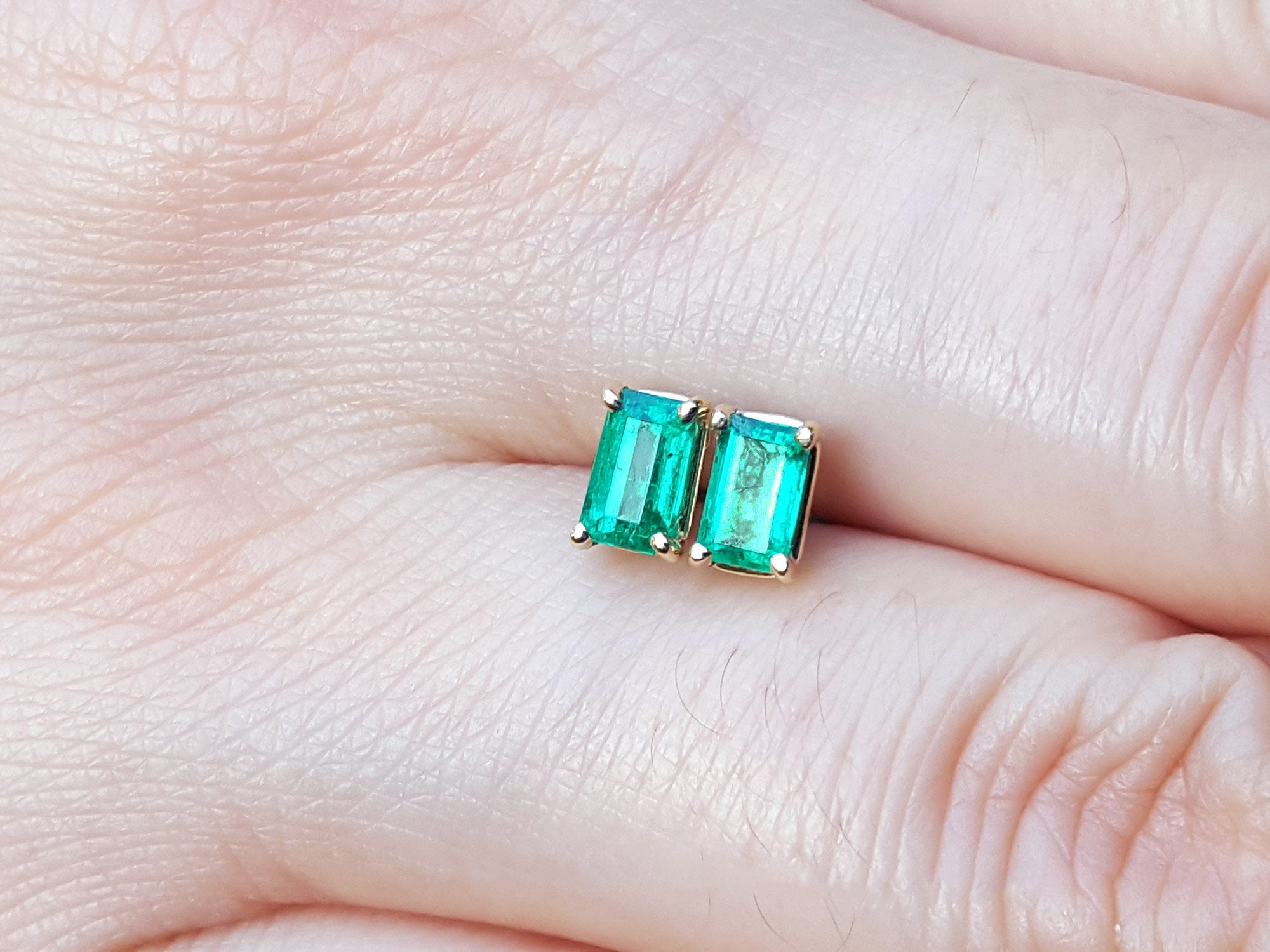 Yellow gold emerald solitaire stud earrings between a person&#39;s finger.
