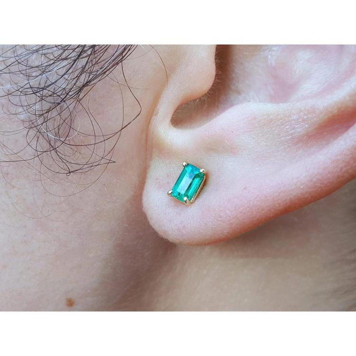An ear wearing a Colombian emerald solitaire stud earring in 14k gold.