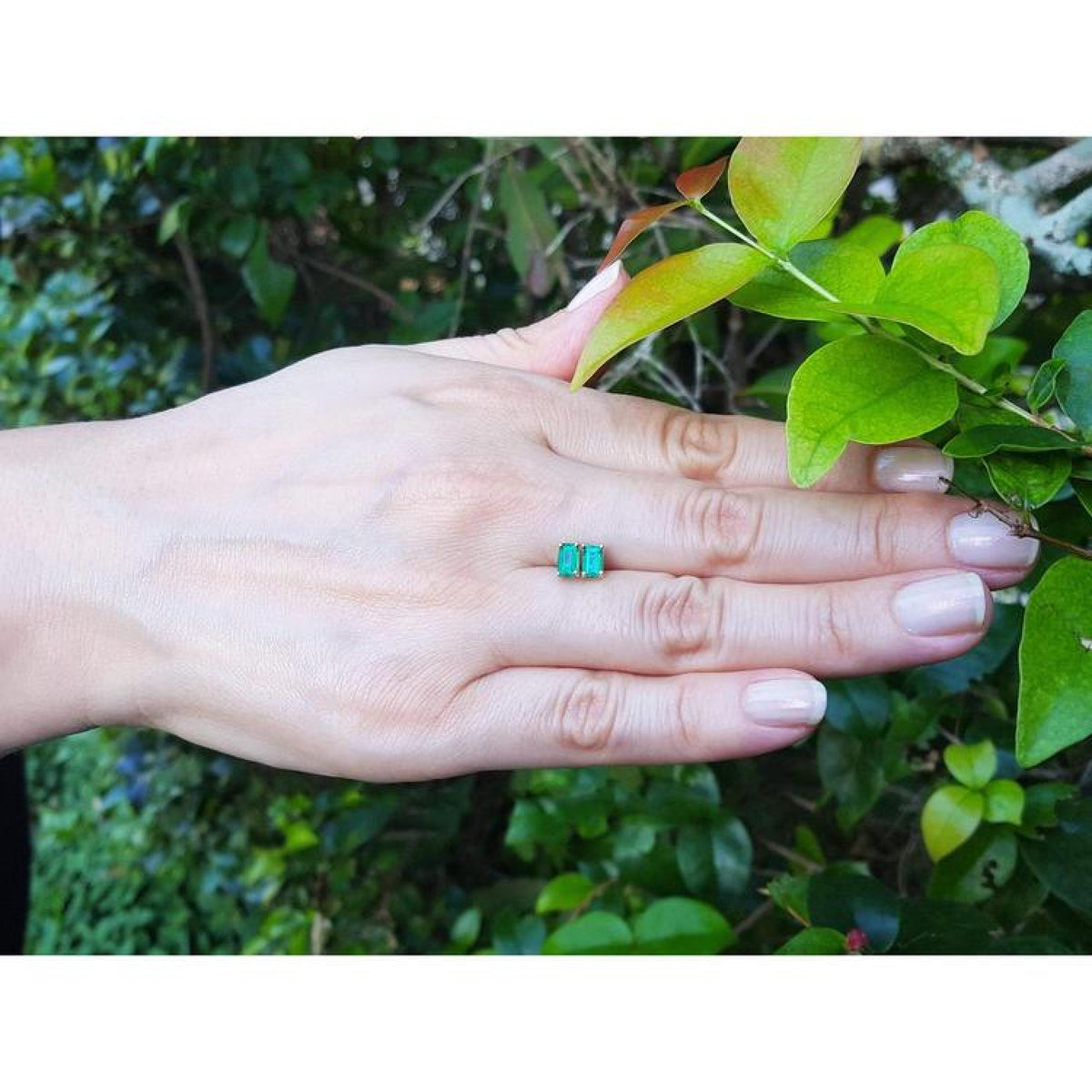 Pair of emerald cut emerald stud earrings in 14K yellow gold, displayed on a hand.