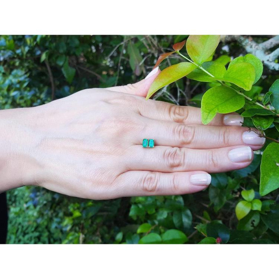 Pair of emerald cut emerald stud earrings in 14K yellow gold, displayed on a hand.