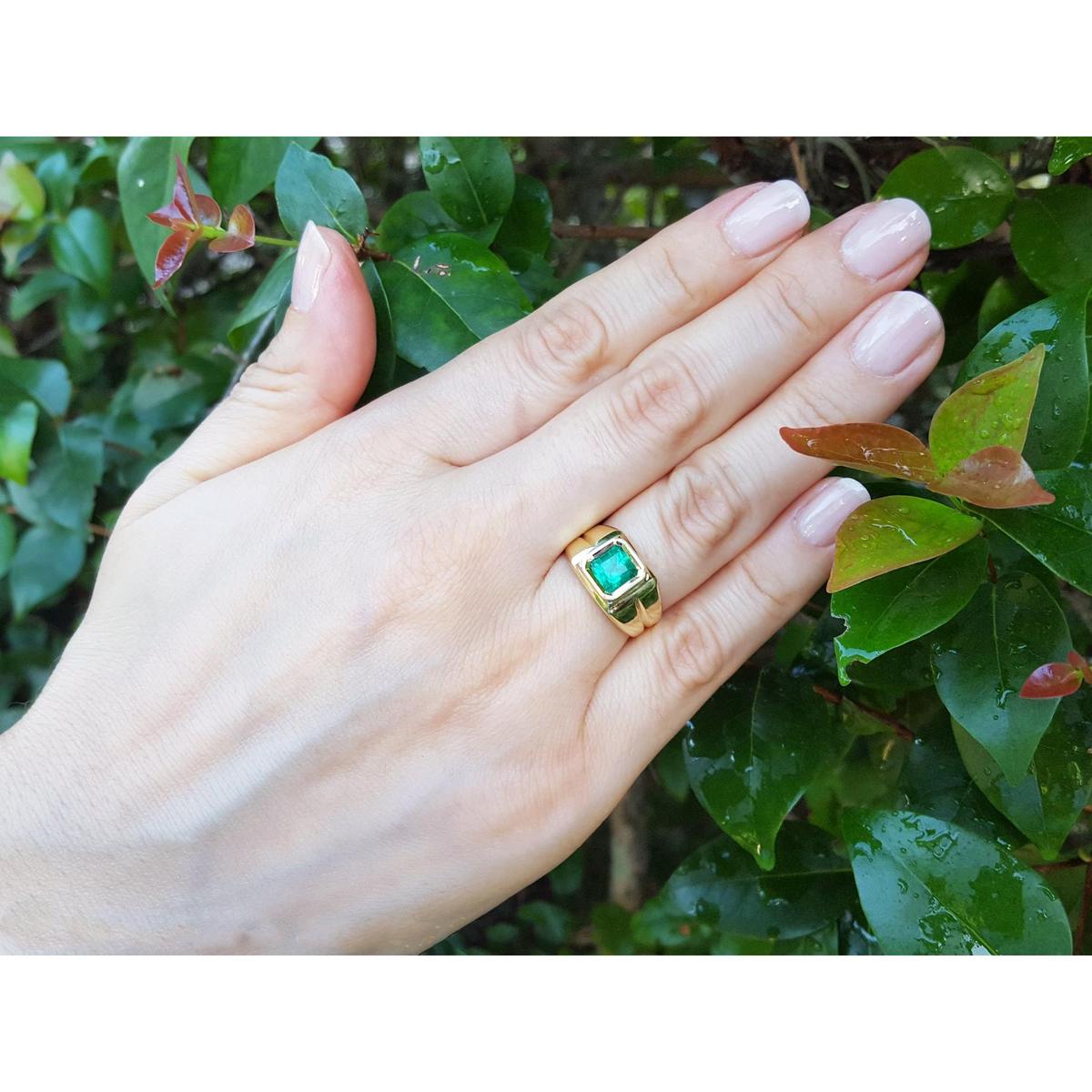 Hand wearing a gold ring with a square genuine colombian emerald against a leafy background