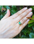 Hand wearing a gold ring with a square genuine colombian emerald against a leafy background