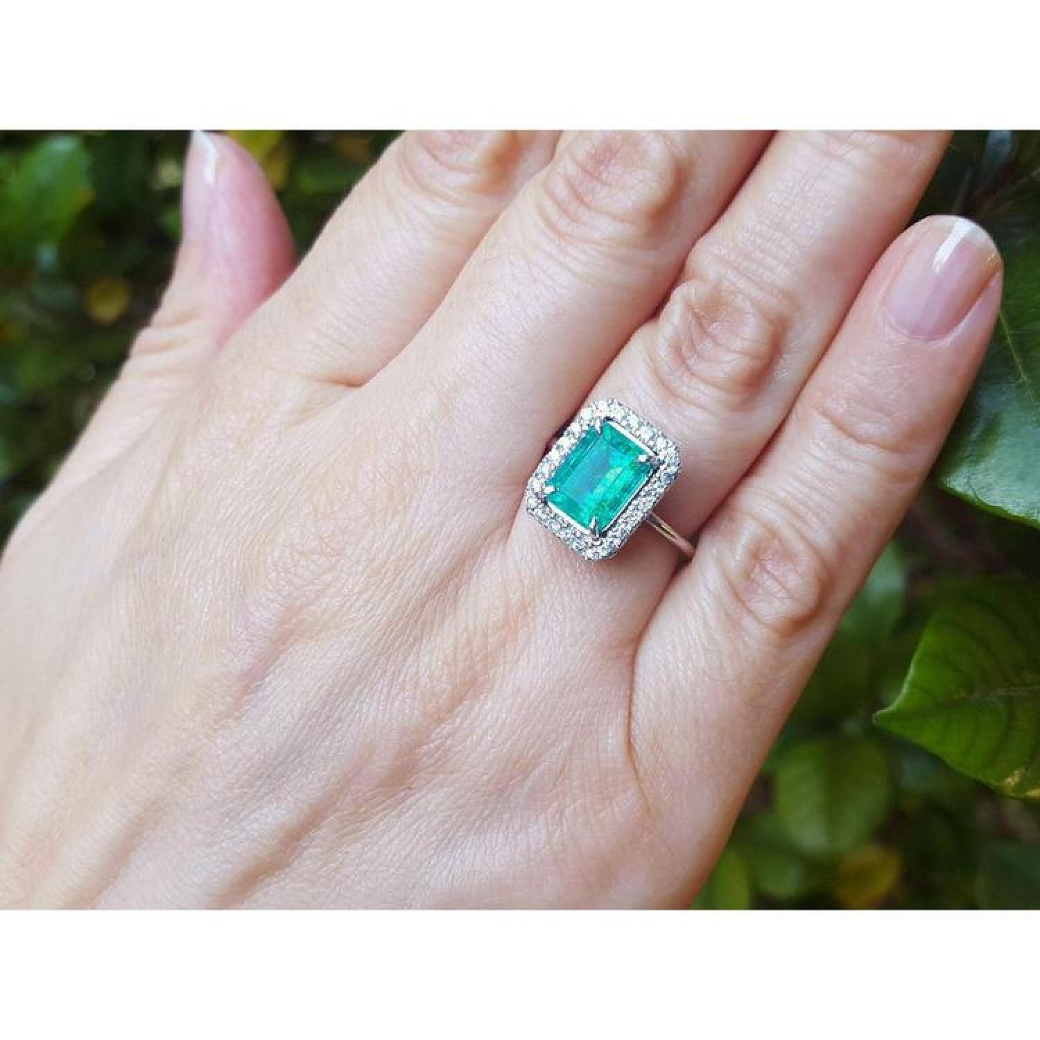 Emerald-cut bluish-green emerald ring with halo diamonds in 14K white gold on a woman's hand.