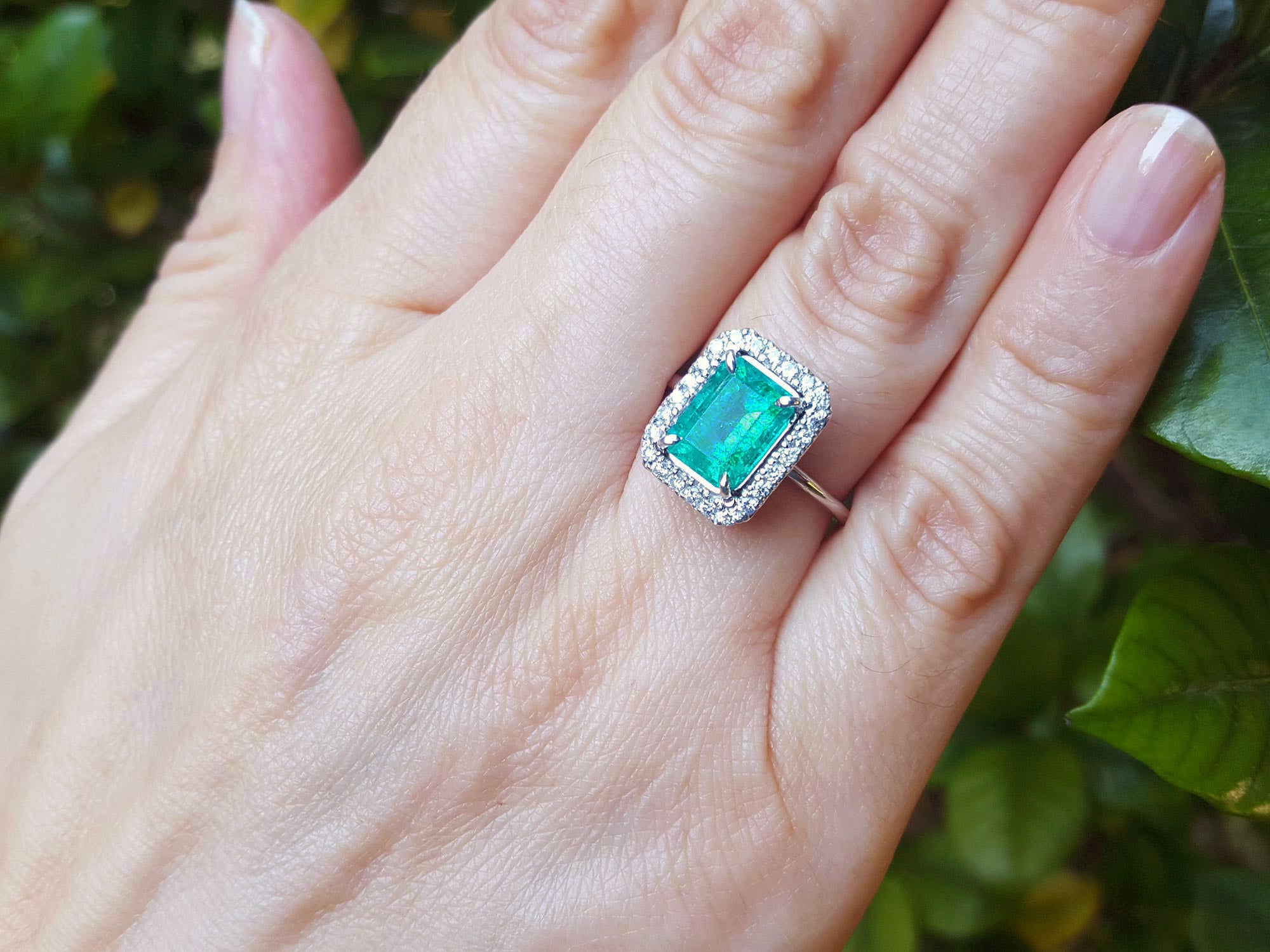 Hand wearing a square emerald ring with a halo diamond against a green leafy background.